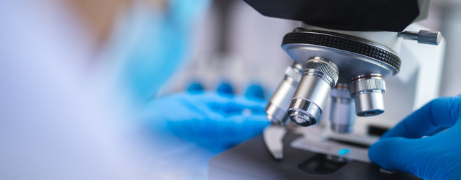 Um microscópio é mostrado em destaque, com um par de mãos usando luvas azuis ajustando a lente. O ambiente sugere um laboratório, com elementos desfocados ao fundo, indicando uma atividade de pesquisa ou análise científica.