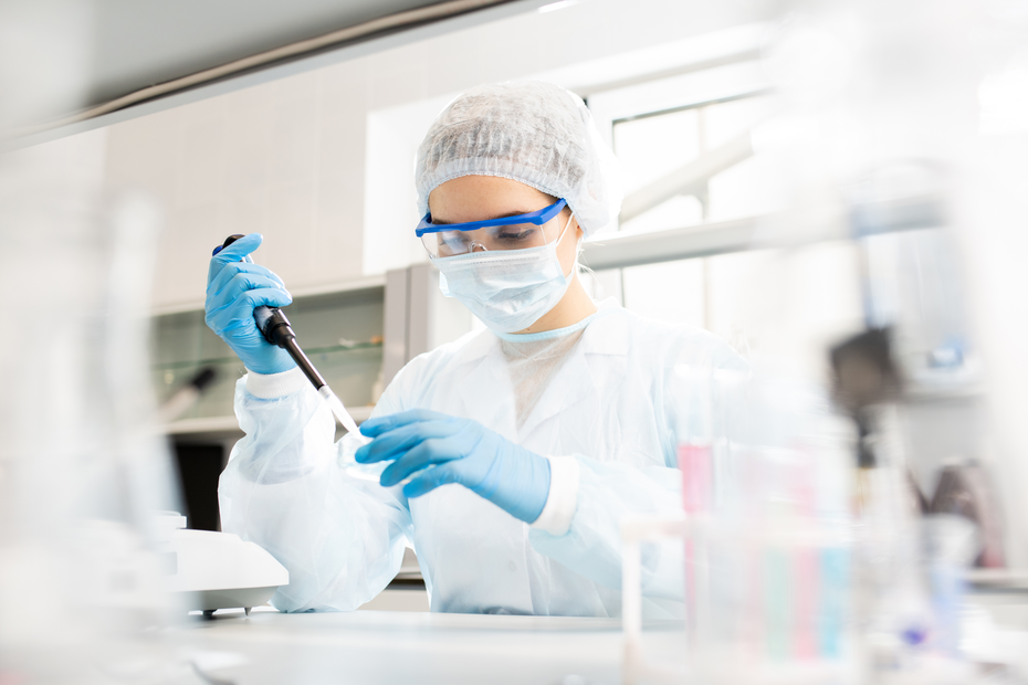 Cientista em um laboratório, usando um jaleco branco, óculos de proteção, touca e máscara. Ela segura uma micropipeta, concentrada em suas atividades. É um ambiente de pesquisa com equipamentos e frascos visíveis ao fundo.