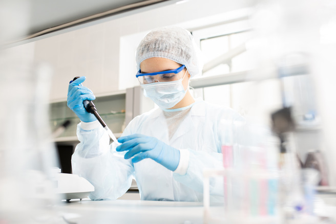 Cientista em um laboratório, usando um jaleco branco, óculos de proteção, touca e máscara. Ela segura uma micropipeta, concentrada em suas atividades. É um ambiente de pesquisa com equipamentos e frascos visíveis ao fundo.
