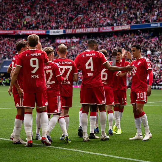 Jogadores do FC Bayern em um momento de confraternização em campo, vestidos com uniformes vermelhos. Alguns jogadores estão se cumprimentando e celebrando, enquanto a torcida ao fundo está animada.