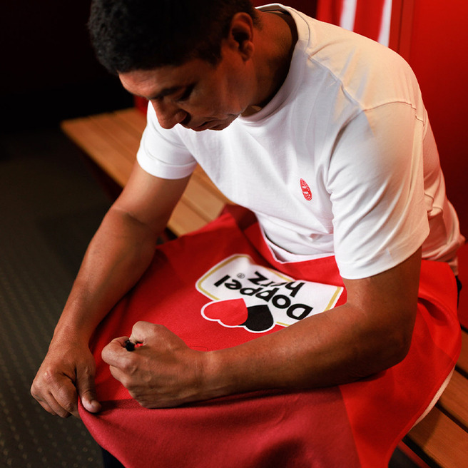 Um homem sentado em um vestiário veste uma camiseta branca e segura uma toalha vermelha com um logo colorido. Ele parece estar ajustando a toalha enquanto se concentra em sua tarefa, com um fundo de assentos de madeira.