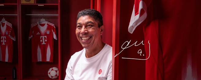 Imagem de um homem sorridente em um vestiário, com paredes vermelhas. Ele está usando uma camiseta branca e há camisas de futebol do Bayern de Munique ao fundo. Uma bola de futebol está visível, e há uma assinatura ao lado dele.