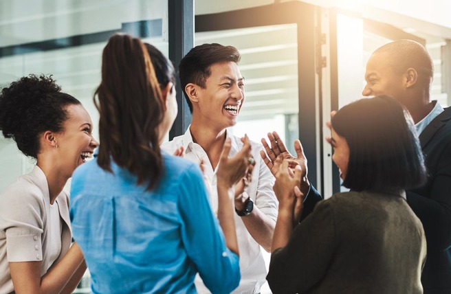 Um grupo de seis pessoas, de diversas etnias, se reúne em um ambiente moderno, sorrindo e aplaudindo. Elas demonstram alegria e camaradagem enquanto conversam animadamente. A luz do sol entra por grandes janelas, criando um ambiente acolhedor.
