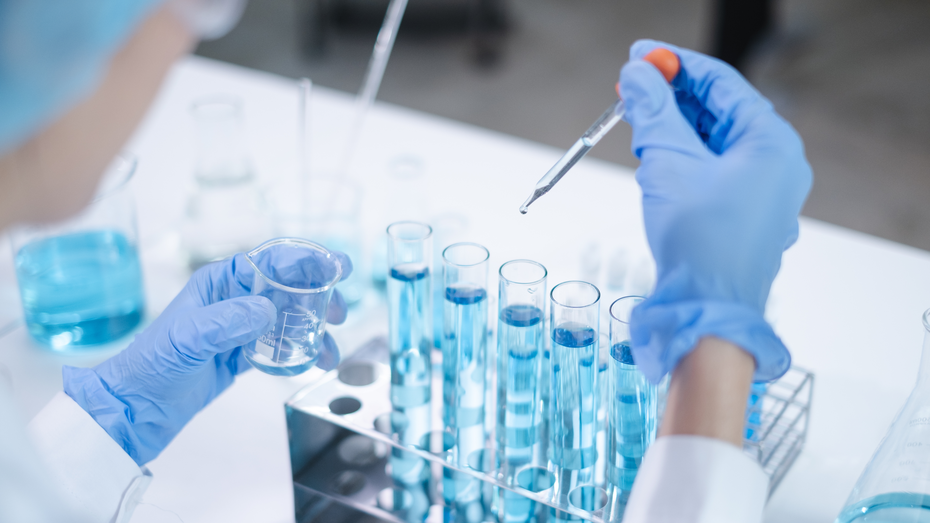 Uma mão com luva azul segura um pequeno béquer enquanto a outra usa uma pipeta para transferir líquido azul para tubos de ensaio em um suporte. O fundo mostra frascos e equipamentos de laboratório.