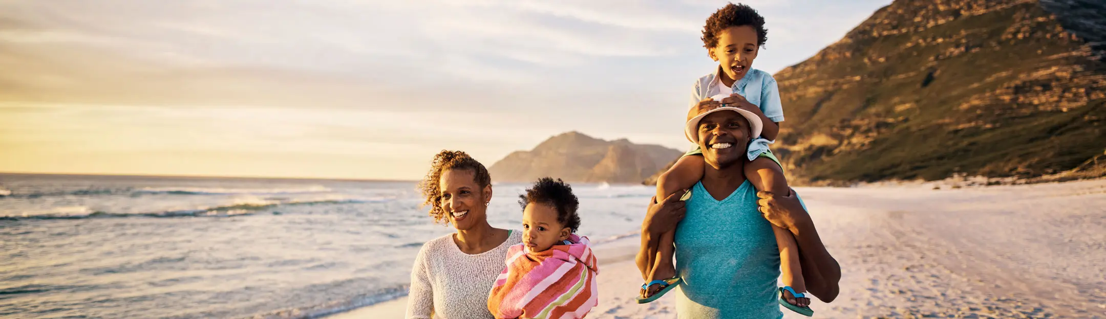 Família na praia durante o pôr do sol, composta por quatro membros: uma mulher com uma criança no colo, uma mulher ao lado e um homem com uma criança nos ombros. Ao fundo, montanhas e mar.