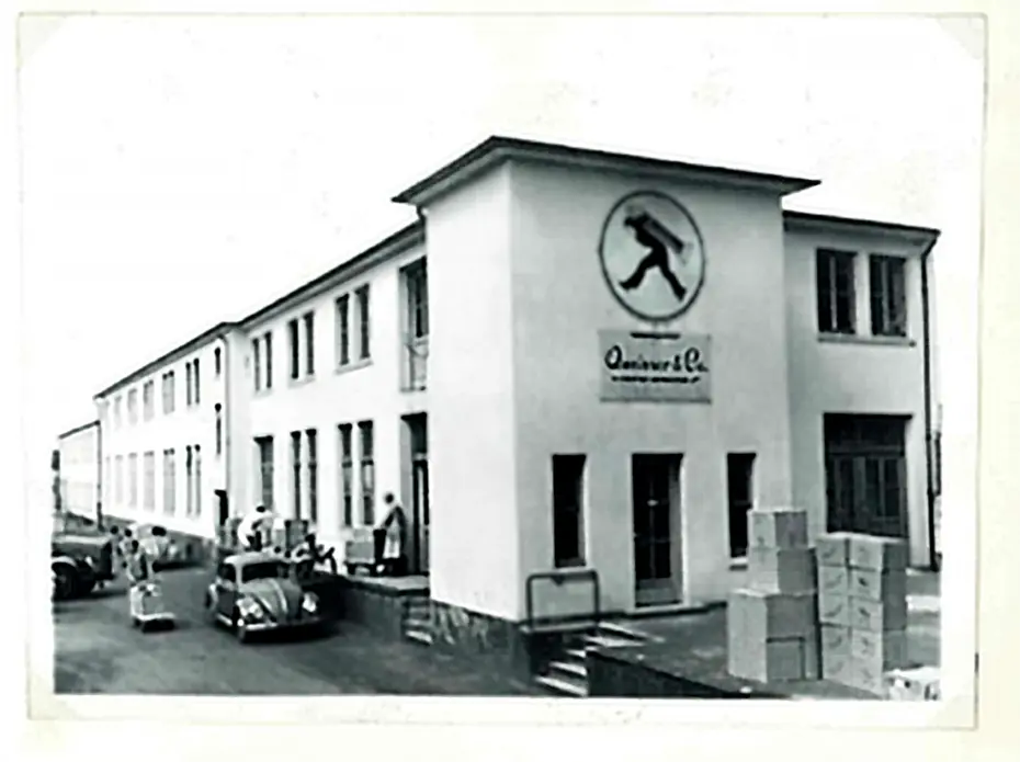 Fachada de um edifício industrial branco com um símbolo de uma pessoa caminhando. Há caixas empilhadas na entrada e algumas pessoas e carros na calçada. O ambiente é urbano e o estilo da foto sugere uma época passada.