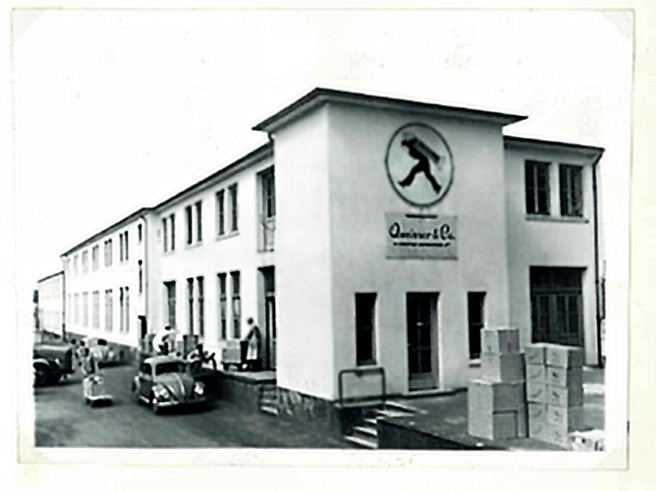 Fachada de um edifício industrial branco com um símbolo de uma pessoa caminhando. Há caixas empilhadas na entrada e algumas pessoas e carros na calçada. O ambiente é urbano e o estilo da foto sugere uma época passada.