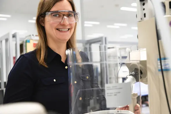 Mulher sorridente usando óculos de segurança em um laboratório. Ela está perto de um equipamento, com uma bancada de trabalho ao fundo. O ambiente parece moderno e bem iluminado, sugerindo um espaço dedicado à pesquisa científica.