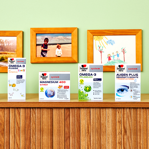 Uma prateleira de madeira exibe produtos de saúde da marca Doppelherz, incluindo suplementos de Omega-3, magnésio e proteção ocular. Ao fundo, há paredes em tom verde com molduras que contêm fotos e desenhos infantis.