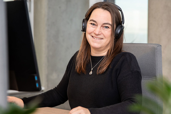 Mulher sorridente usando um headset, sentada em frente a um computador. Ela tem cabelo longo e castanho e está vestindo um suéter escuro. O ambiente parece um escritório moderno.