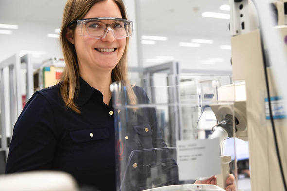 Mulher sorridente usando óculos de segurança em um laboratório. Ela está perto de um equipamento, com uma bancada de trabalho ao fundo. O ambiente parece moderno e bem iluminado, sugerindo um espaço dedicado à pesquisa científica.
