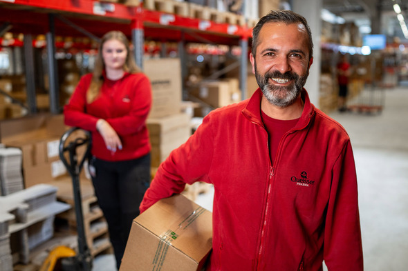 Dois trabalhadores em um ambiente de armazém. Um homem sorri enquanto segura uma caixa, vestindo uma camiseta vermelha. Ao fundo, uma mulher também vestindo vermelho está próxima a uma empilhadeira, observando. O local é organizado, com prateleiras cheias de caixas.