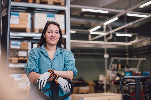 Mulher sorridente vestindo uma camisa azul e luvas de segurança, apoiando-se em um equipamento de movimentação de carga. Ao fundo, prateleiras com caixas e um ambiente de trabalho industrial.